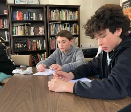 Two boys sit at a table, writing on sheets of paper, with bookshelves filled with books in the background.