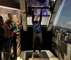 A person hangs from a horizontal bar in a museum exhibit while several people watch from the side.