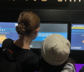 Two people, one with a braided bun and one wearing a white knit hat, stand in front of digital screens in a dark indoor setting.