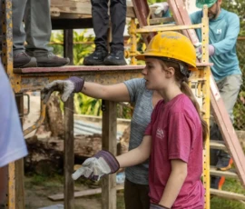 Several people wearing gloves and hard hats work together on a construction site, standing near scaffolding and a ladder outdoors.