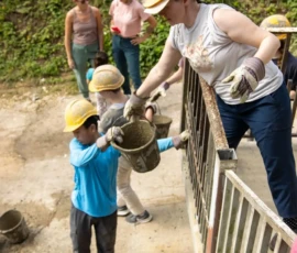 A woman wearing a hard hat and gloves passes a bucket to a child in a construction area, while others with helmets stand nearby outdoors.