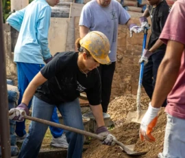 Five people wearing gloves and hard hats are shoveling and transferring dirt into buckets at a construction site.