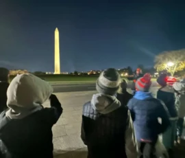 A group of people, some in winter clothing, stand at night facing the illuminated Washington Monument in the distance.